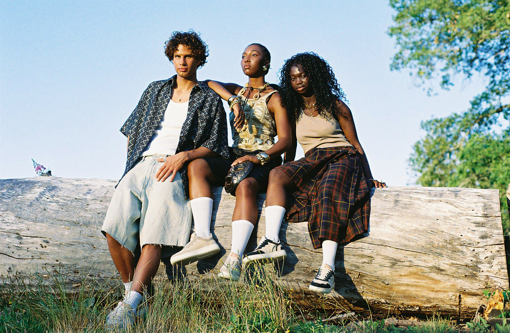 Three people sitting on a log in a natural setting with trees and clear sky.
